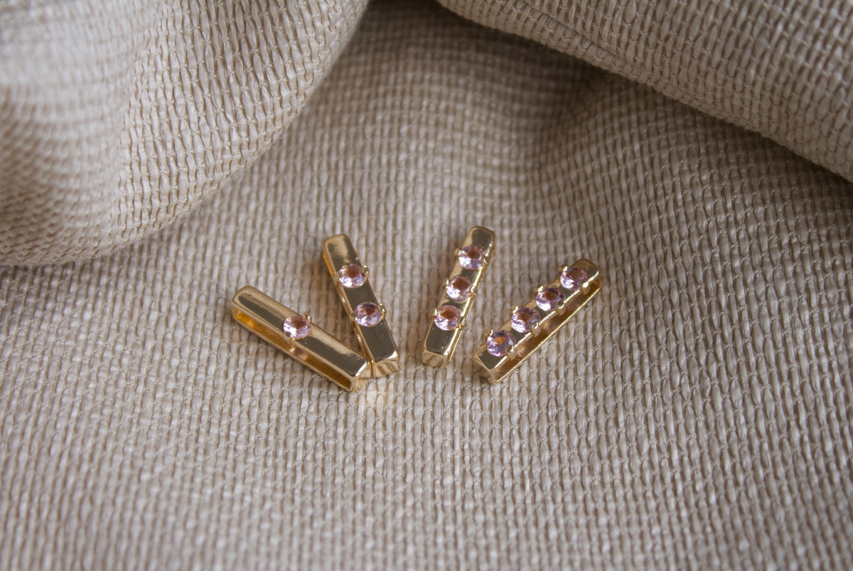 Four gold jewelry sliders, each embedded with sparkling pink cubic zirconia stones, rest on a textured beige fabric.