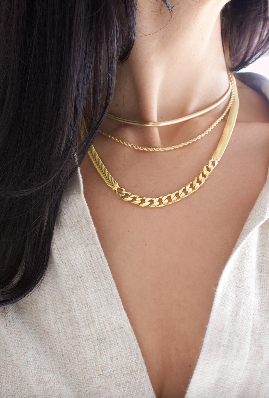 A woman with dark hair wears a layered gold chain necklace set, featuring a smooth snake chain, a delicate rope chain, and a bold herringbone and curb chain combination.