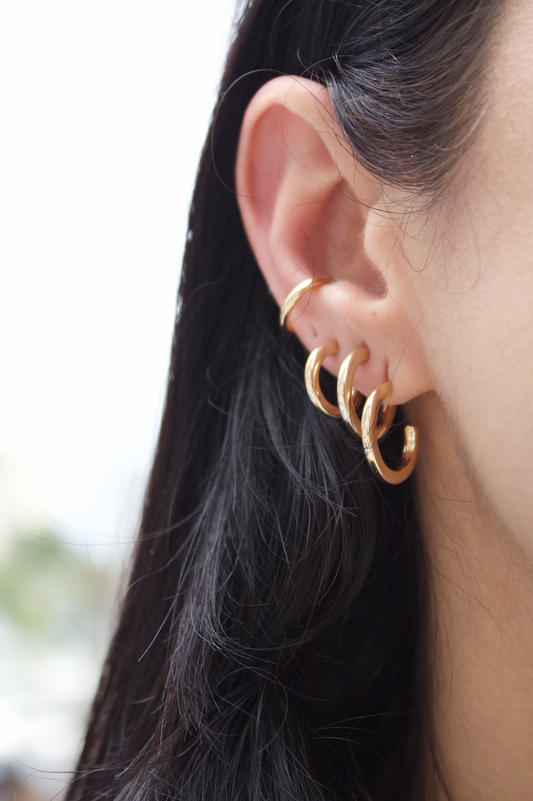 A woman with dark hair showcases a curated ear stack featuring multiple gold hoop earrings and a matching gold ear cuff.