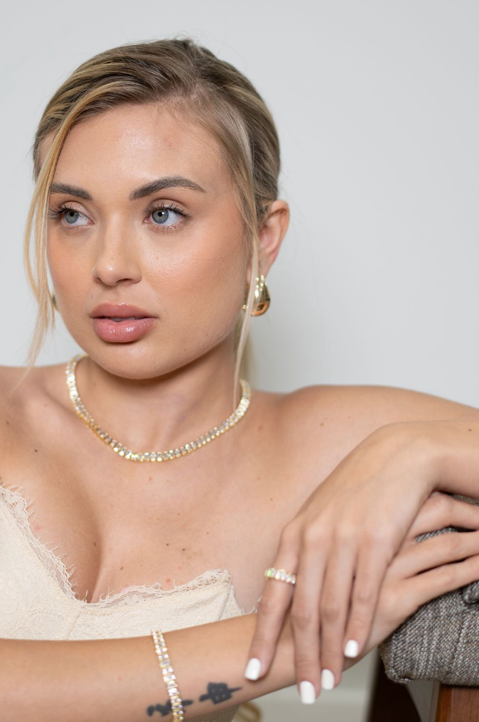 A blond woman wearing a sparkling tennis necklace, matching bracelet, ring, and gold earrings poses thoughtfully.