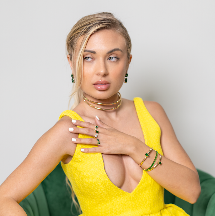 A blonde woman wearing a vibrant yellow dress showcases luxurious gold jewelry featuring green emerald gemstones, including rings, a choker, and bracelets.