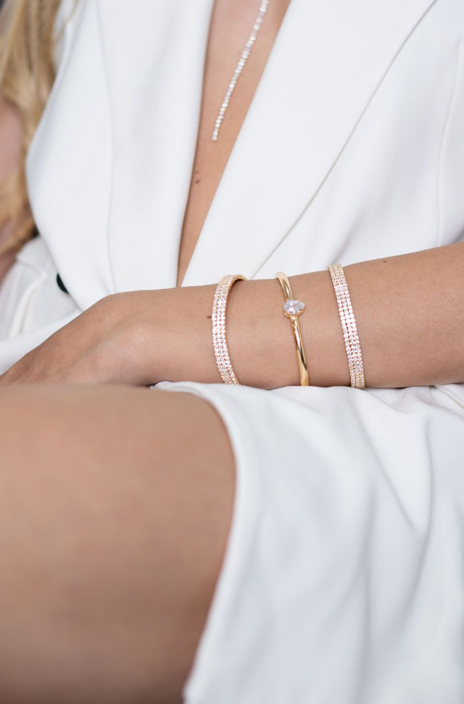 A woman wearing a crisp white blazer showcases a stack of sparkling diamond bracelets and a matching tennis bracelet on her wrist.