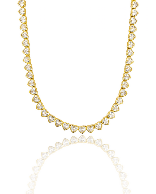 A sparkling gold tennis necklace features a continuous line of heart-cut cubic zirconia stones against a white background.