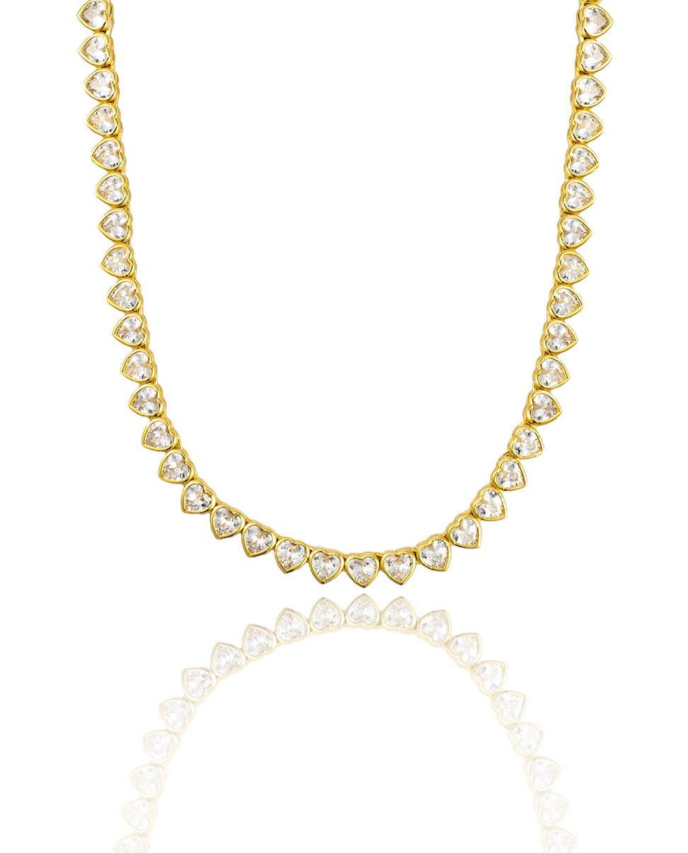 A dazzling gold tennis necklace features a continuous strand of shimmering heart-cut cubic zirconia stones against a clean white background.