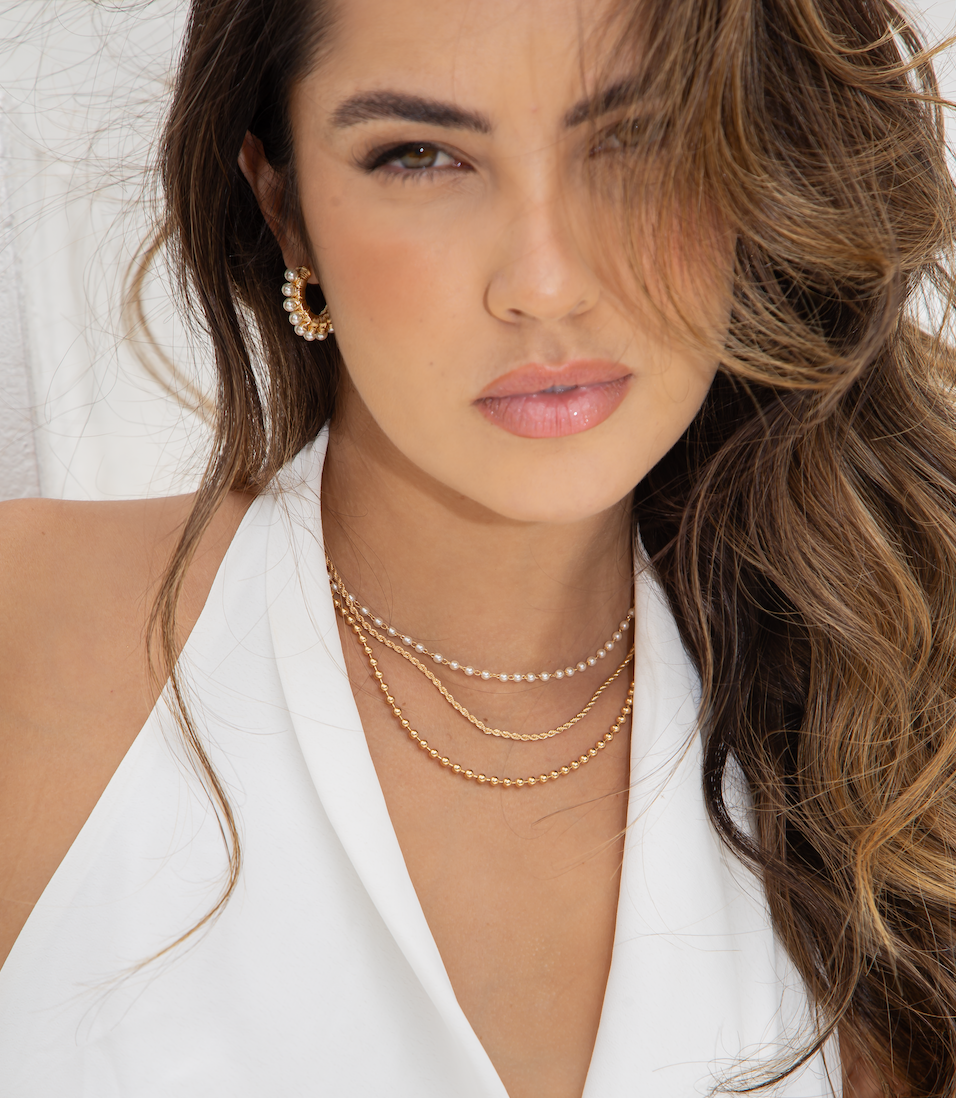 A woman with layered gold necklaces and pearl hoop earrings poses in a white halter top.