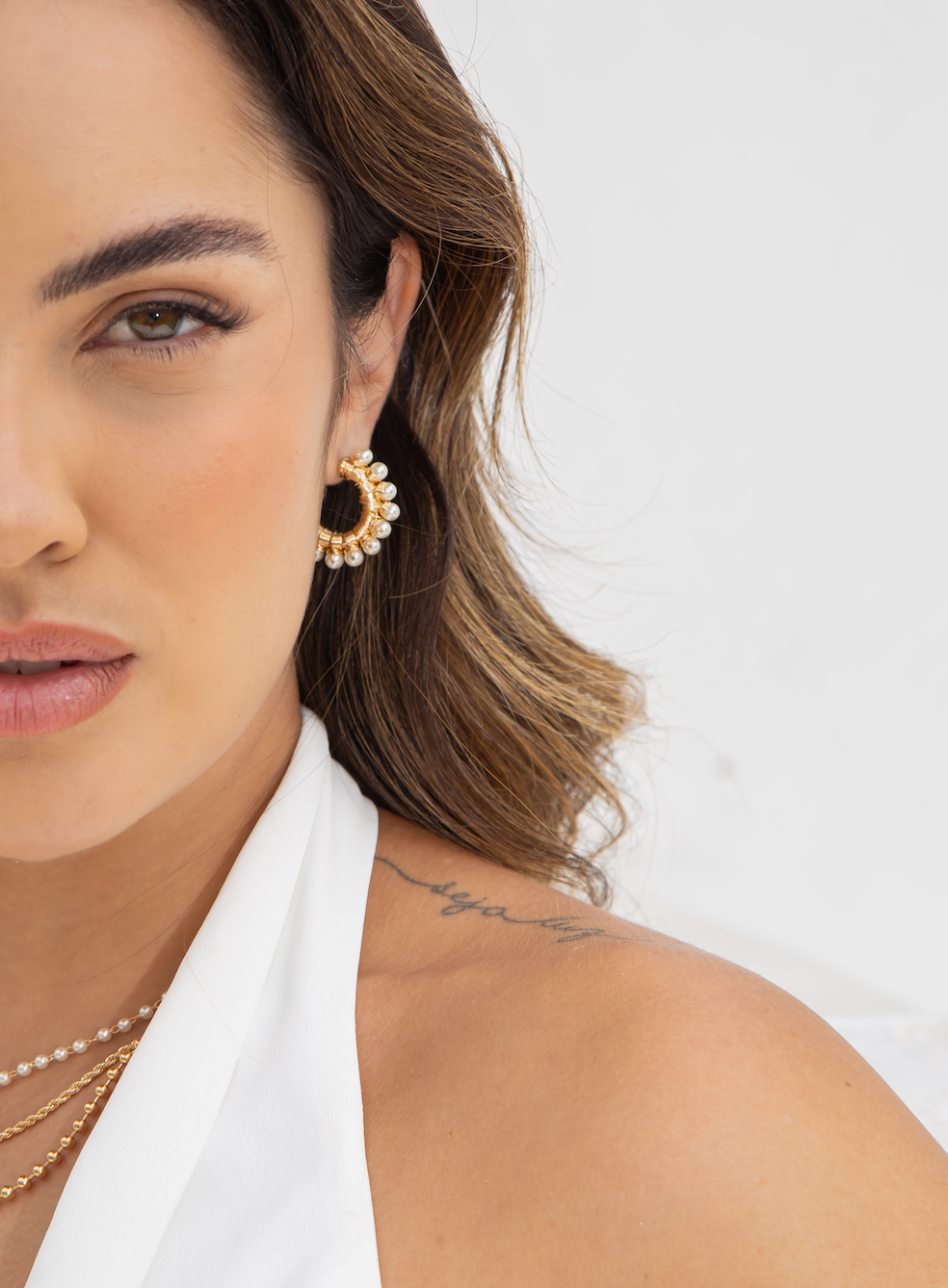 A woman wears a gold hoop earring adorned with small pearls and delicate layered necklaces.