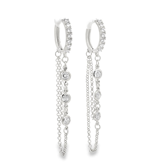 A pair of sterling silver huggie hoop earrings features sparkling pavé-set diamonds and delicate dangling chains adorned with bezel-set stones against a clean white background.