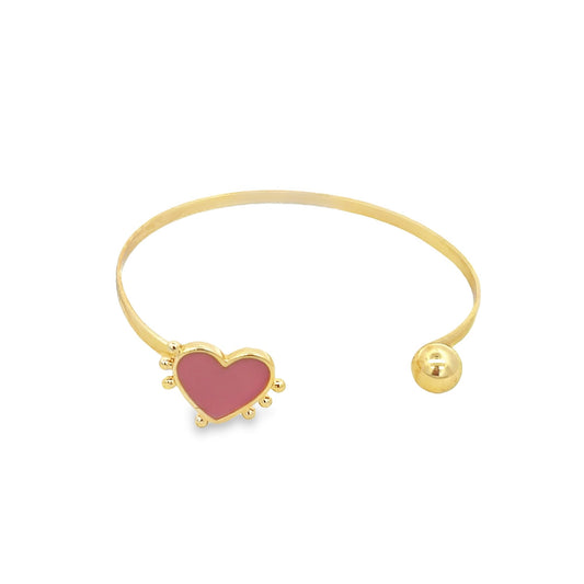 A gold cuff bracelet features a pink enamel heart charm on one end and a polished ball on the other against a white background.
