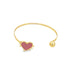 A gold cuff bracelet features a pink enamel heart charm on one end and a polished ball on the other against a white background.