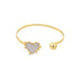 A gold cuff bracelet features a mother-of-pearl heart charm on one end and a polished gold sphere on the other.