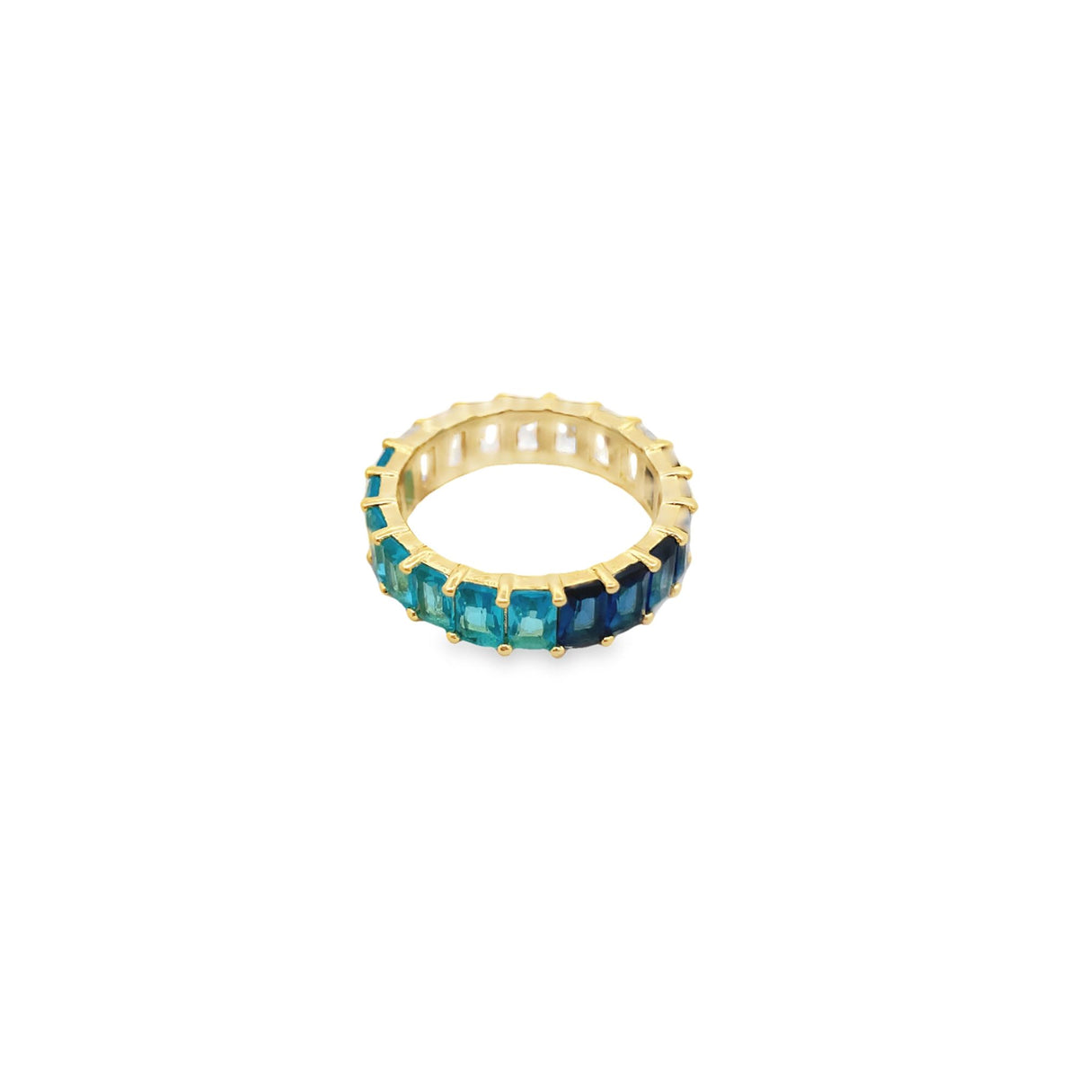 A gold eternity ring features an ombre of baguette-cut blue and teal gemstones against a plain white background.