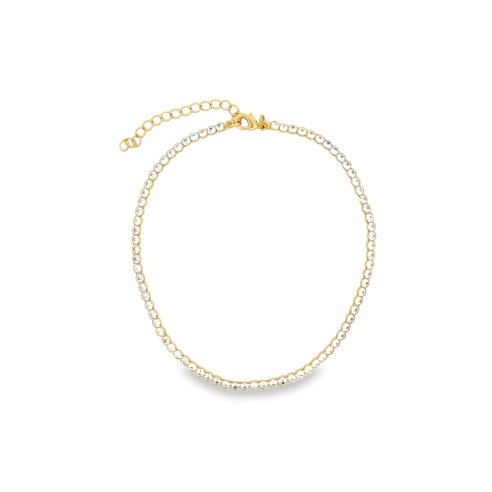A gold tennis necklace featuring a continuous line of sparkling clear rhinestones rests in a circle on a white background.