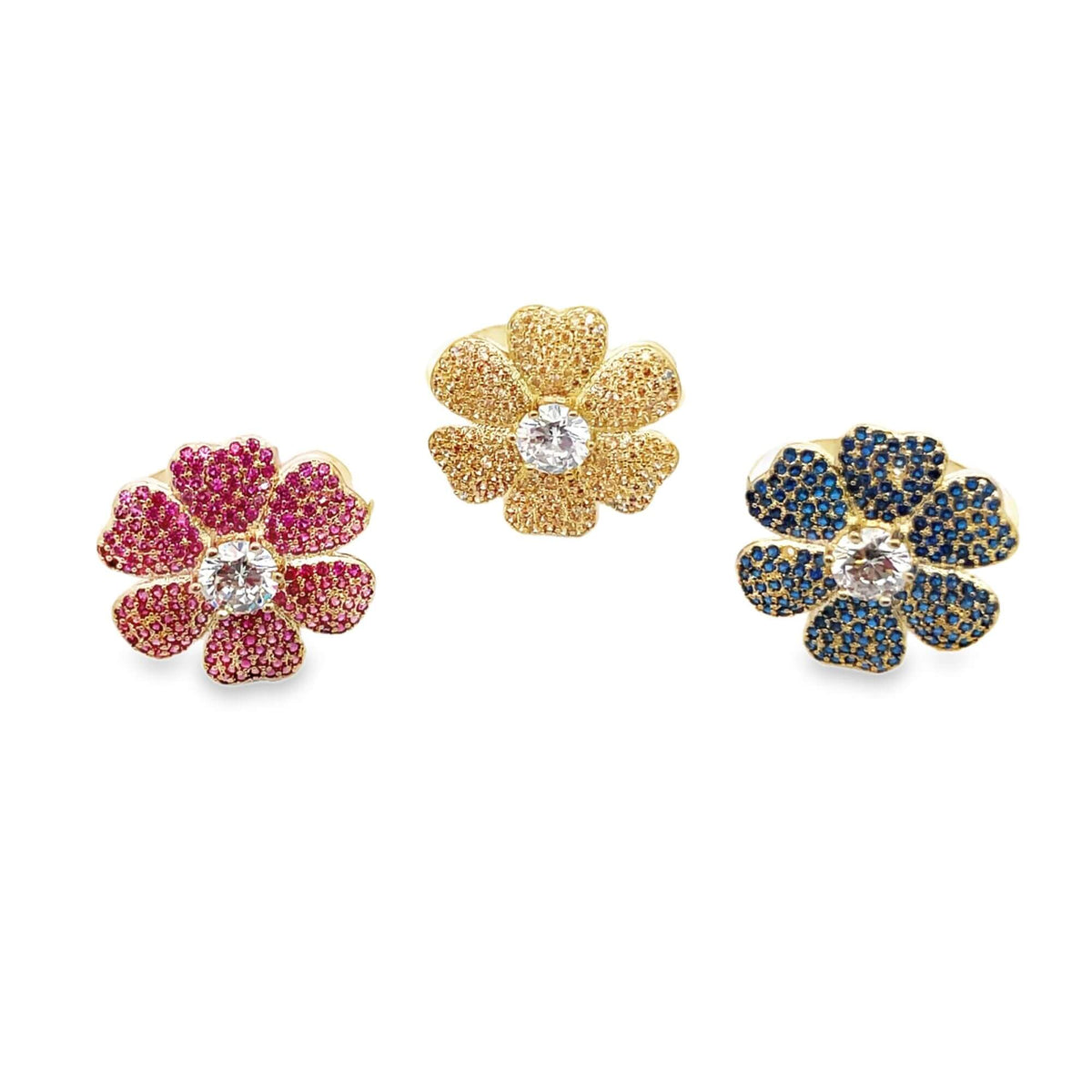 Three sparkling gold flower rings feature pavé-set pink, yellow, and blue gemstones with central diamond-like stones against a white background.