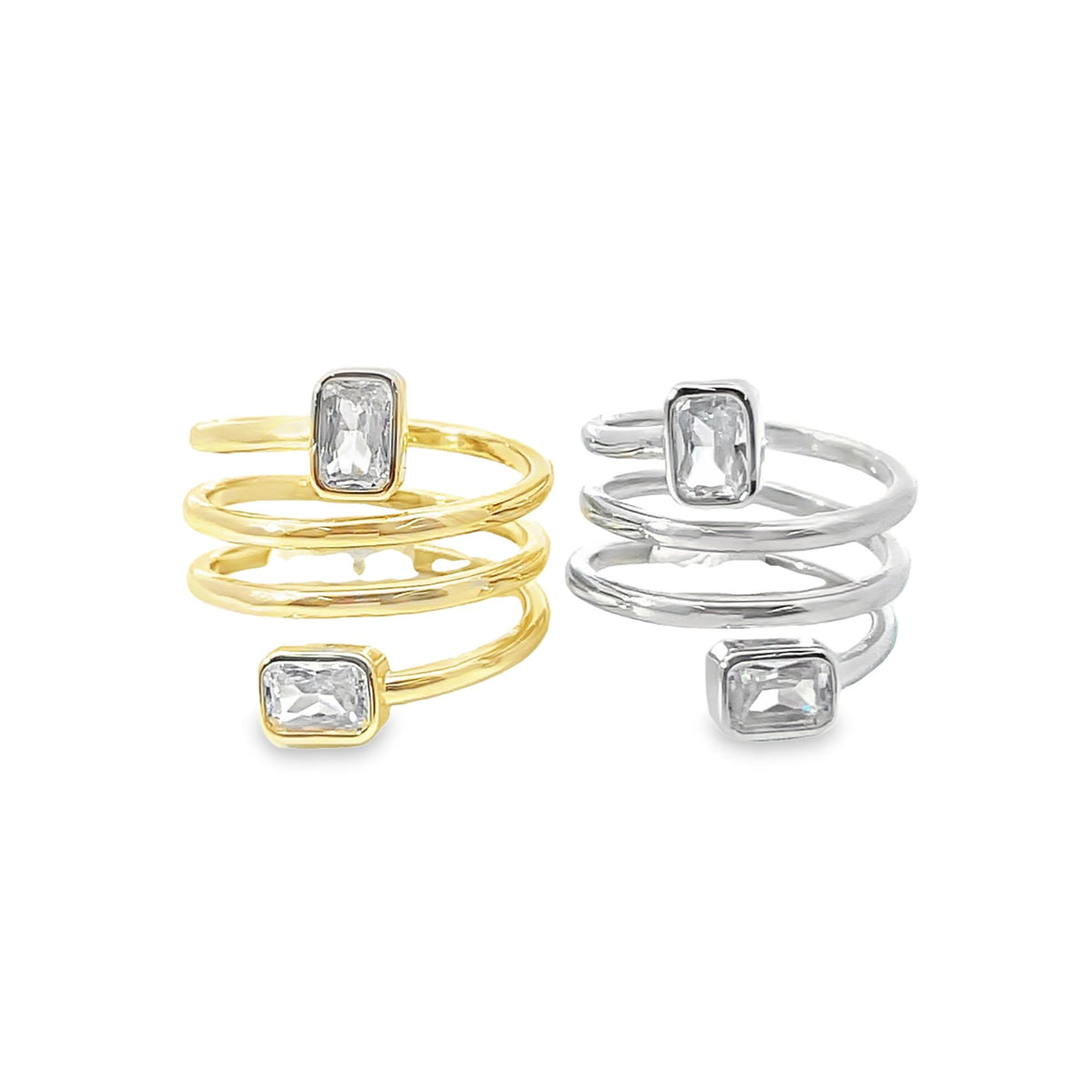 Two spiral bypass rings featuring rectangular cubic zirconia stones rest side by side, one in gold and one in silver.