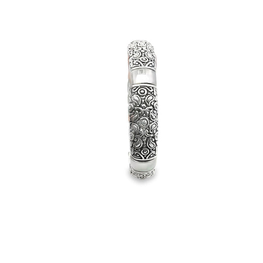 An ornate silver bangle bracelet displays intricate engraved patterns and smooth metal sections against a white background.