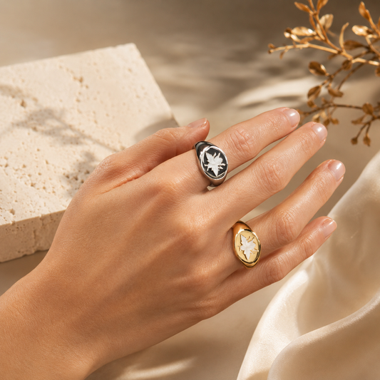 A woman displays a silver signet ring with a black enamel face and a gold signet ring featuring mother-of-pearl floral inlays on her fingers against a neutral textured backdrop.