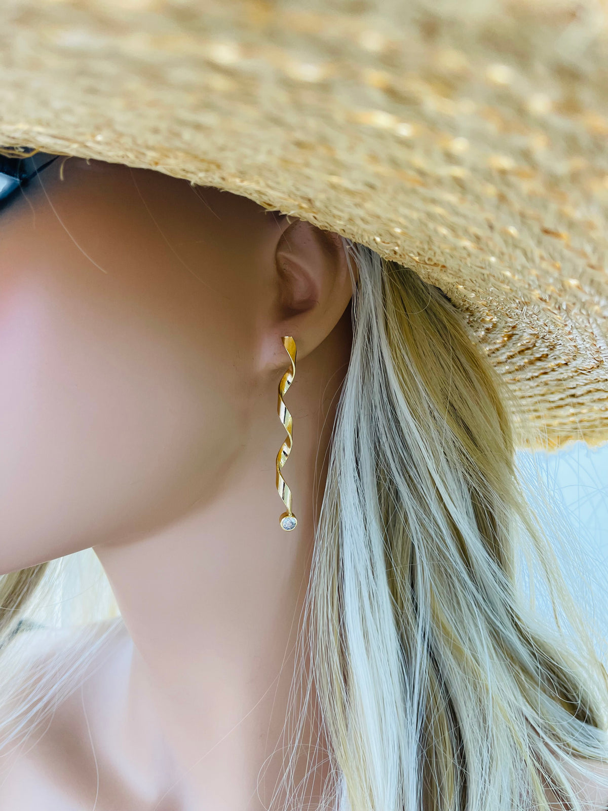 A woman wearing a stylish straw hat showcases a long, gold spiral dangle earring set with a sparkling cubic zirconia stone at the tip.