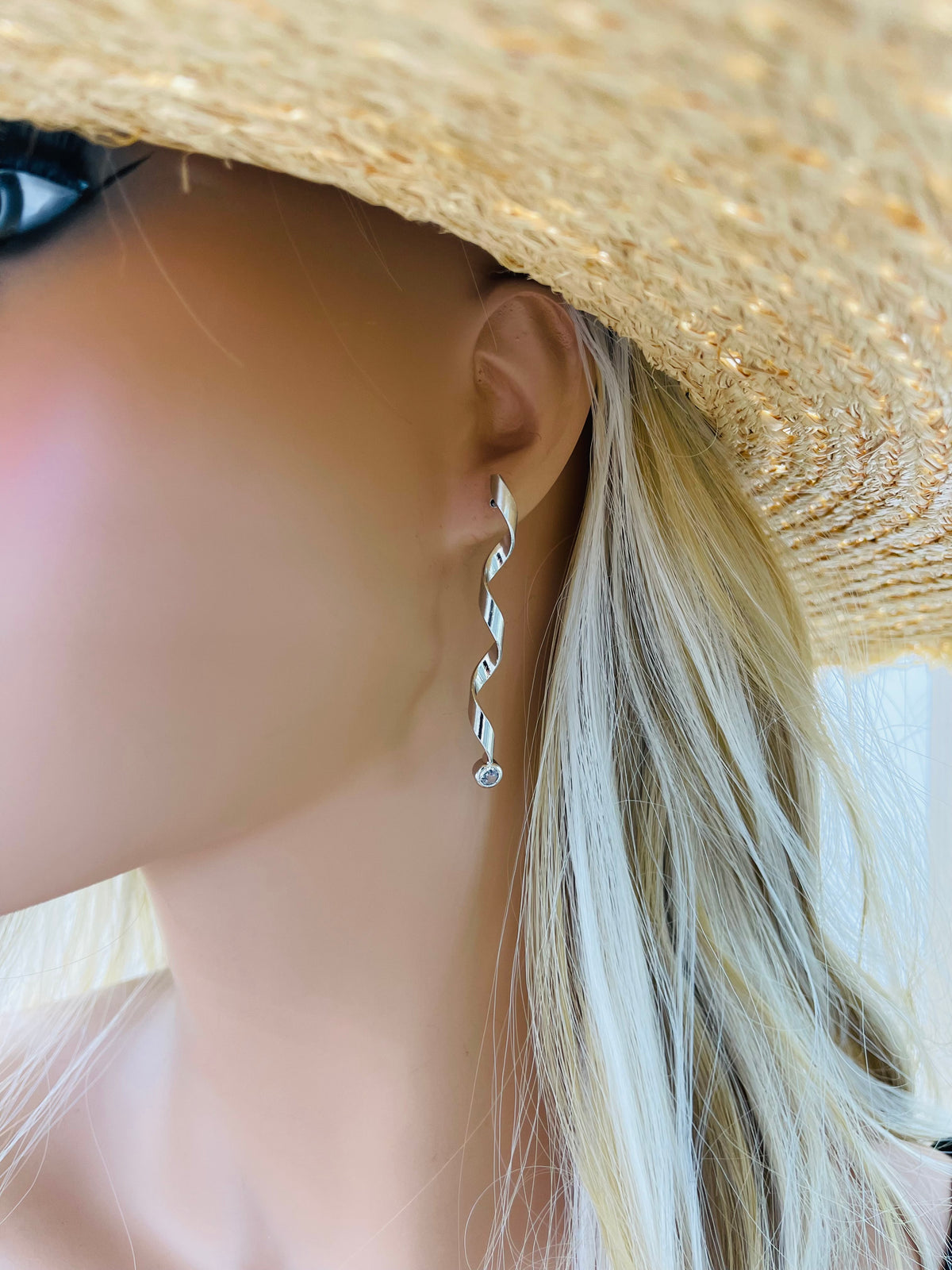 A blonde mannequin wearing a straw hat models a long, spiraling silver dangle earring featuring a small gemstone at the bottom.