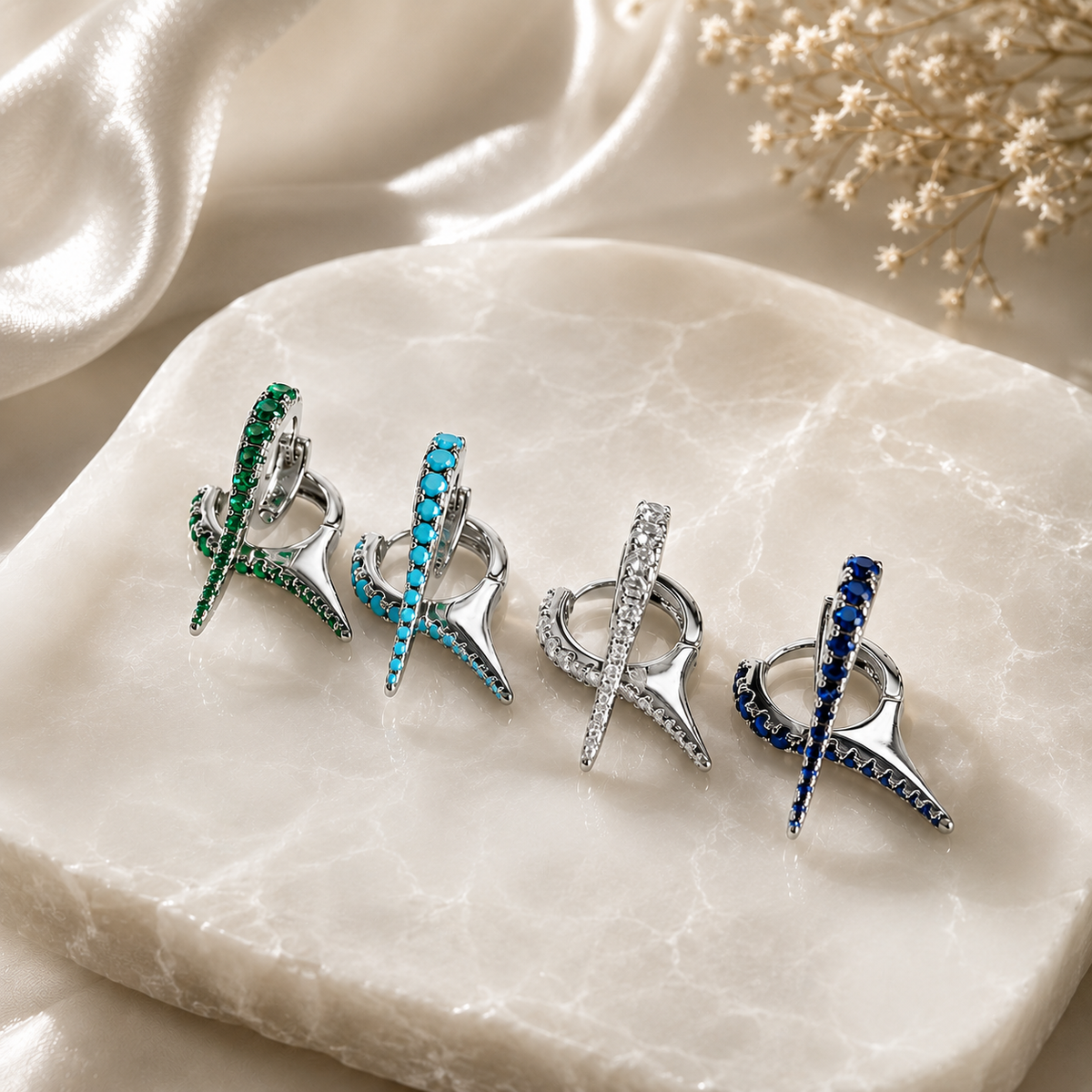 Four pairs of silver huggie hoop earrings featuring dagger-shaped charms studded with green, turquoise, white, and blue crystals rest on a white marble slab.