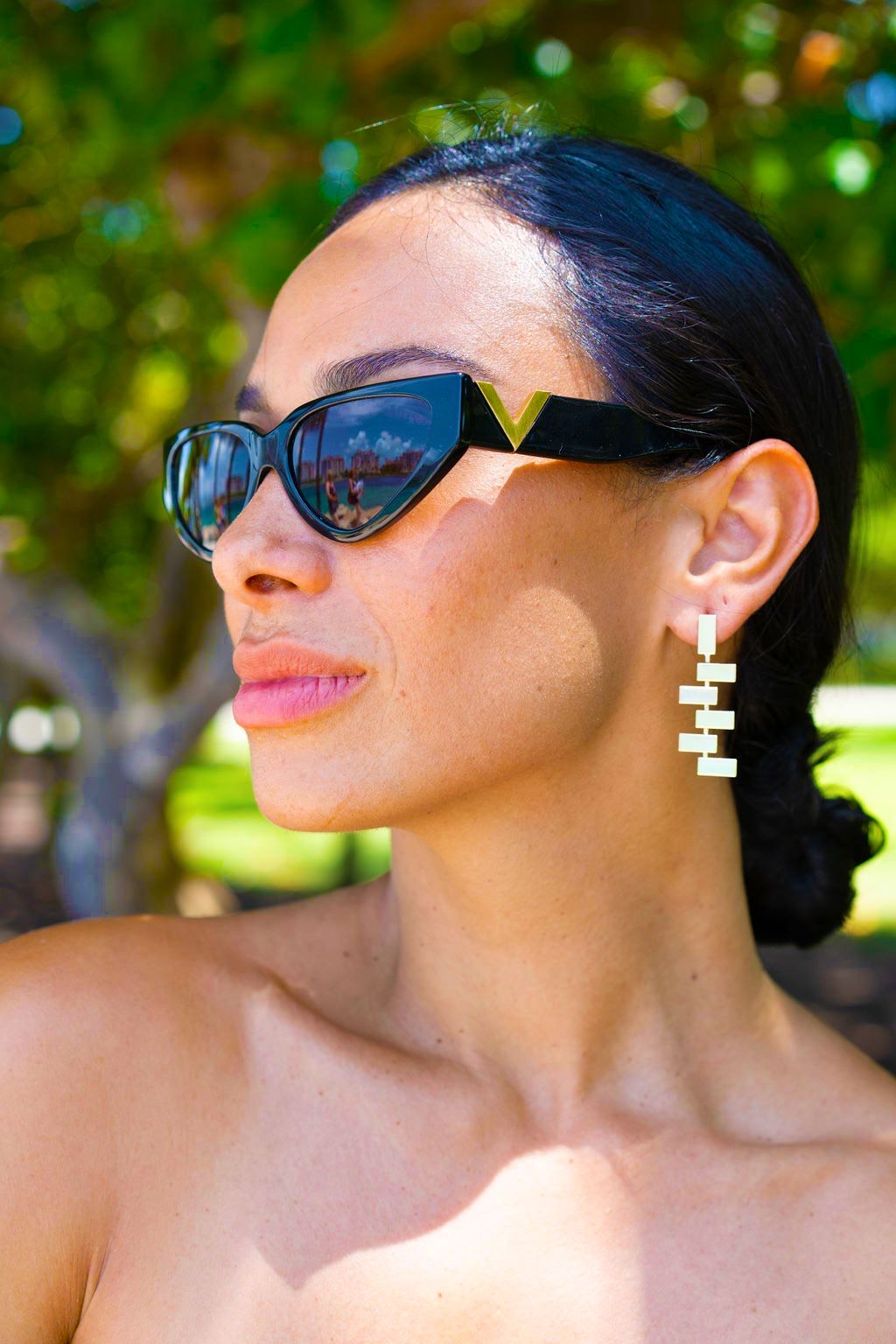 A stylish woman with dark hair wearing black cat-eye sunglasses and geometric gold dangle earrings outdoors in the sunlight.