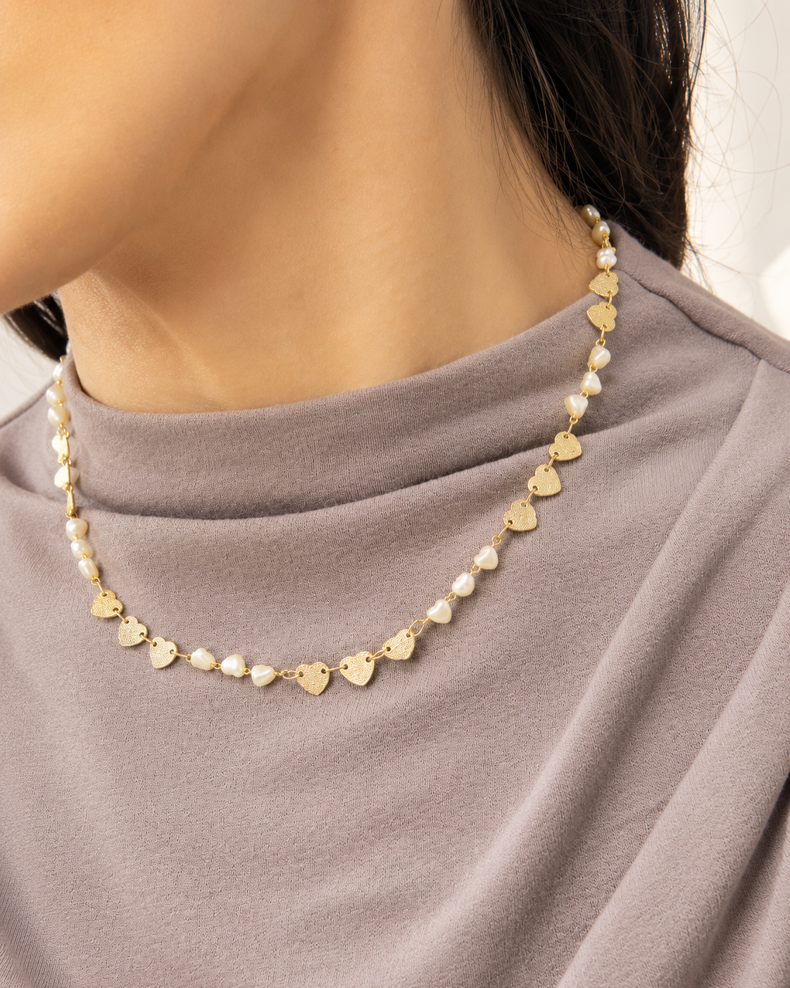 A model wears a delicate gold heart and pearl beaded necklace against a grey top.