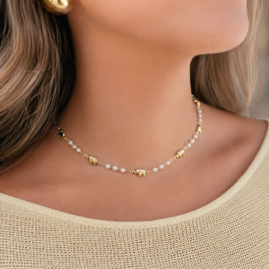 A woman wears a delicate gold chain necklace adorned with small white pearls and tiny gold elephant charms.