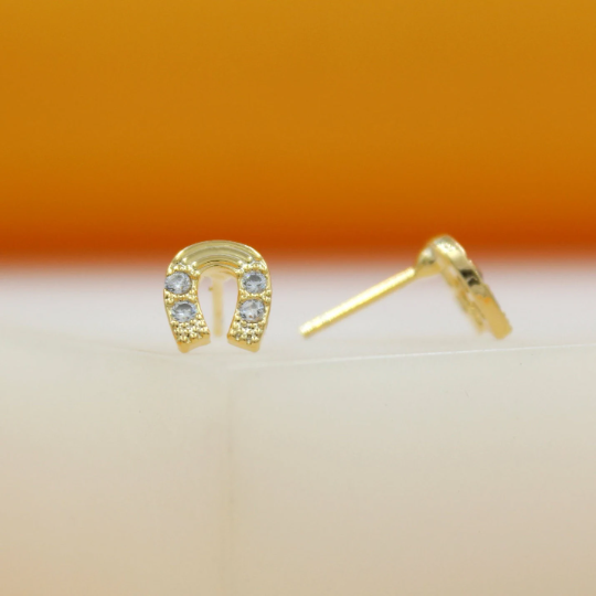 A pair of gold horseshoe stud earrings sparkled with cubic zirconia stones rests upright on a white surface against an orange backdrop.