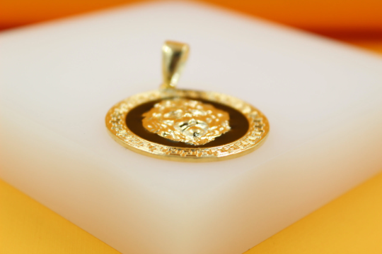 A round gold pendant featuring an embossed lion head against a black enamel background rests on a white display surface.