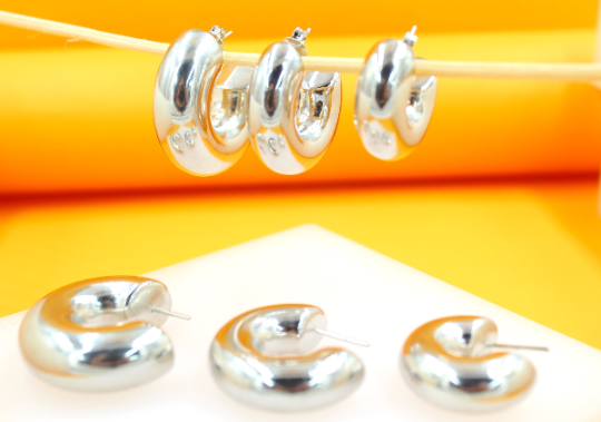 Three pairs of chunky silver hoop earrings in descending sizes rest on a white surface below three matching earrings hanging from a wooden stick against a bright yellow background.