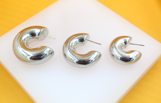 Three graduated silver chunky hoop earrings rest on a white display stand against a vibrant yellow background.