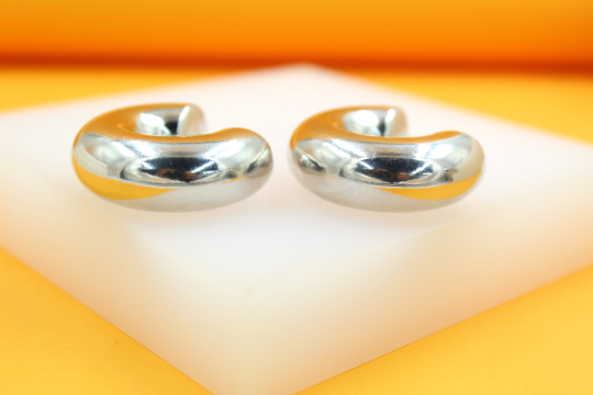 A pair of chunky silver hoop earrings rests on a white display block against an orange backdrop.