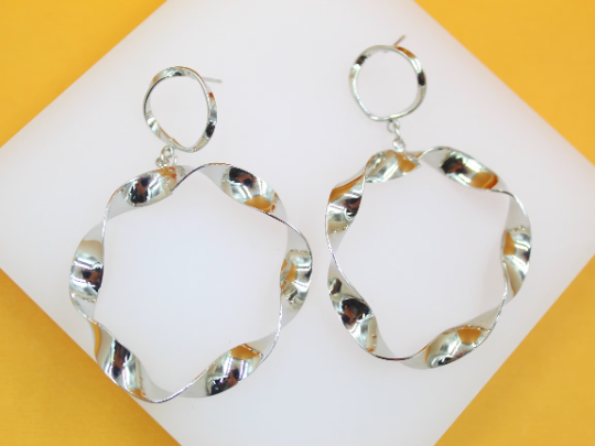 A pair of shiny silver dangle earrings featuring twisted hoop designs rests on a white display surface against a vibrant yellow background.