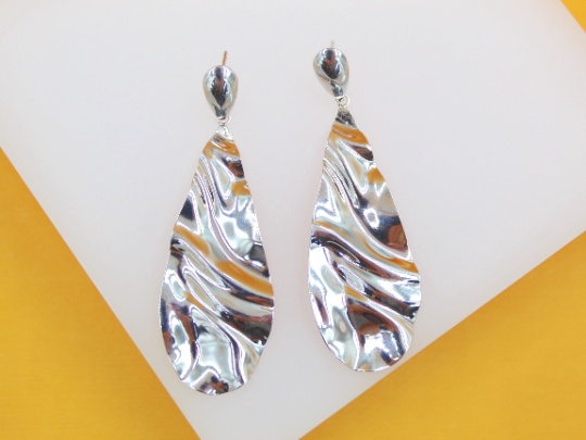 A pair of silver teardrop earrings with a wavy, metallic texture rests on a white display against a yellow background.