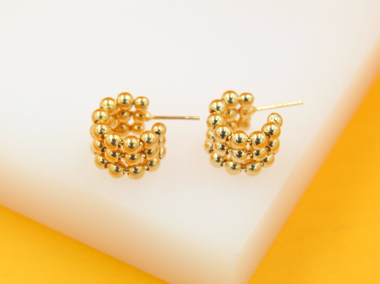 A pair of gold-plated huggie hoop earrings made of tiny metallic beads rests on a white display surface.