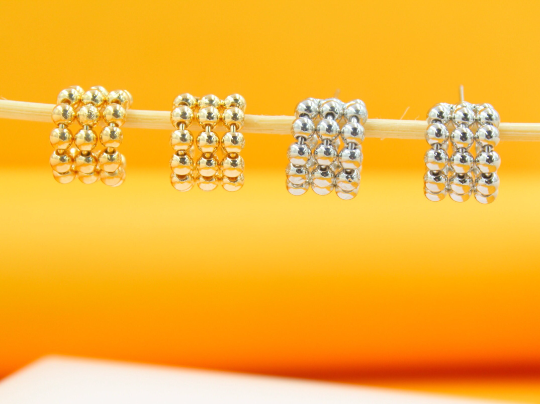Four gold and silver beaded huggie hoop earrings hang delicately on a thin wooden stick against a bright orange background.