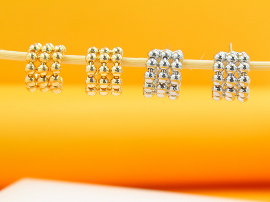Four gold and silver beaded huggie hoop earrings hang delicately on a thin wooden stick against a bright orange background.