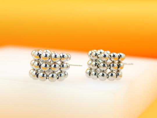 A pair of shiny silver beaded hoop earrings rests on a white display surface against a warm yellow background.