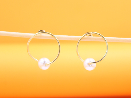 A pair of elegant gold hoop earrings featuring delicate pearl charms rests on a thin wooden stick against a warm, yellow gradient background.