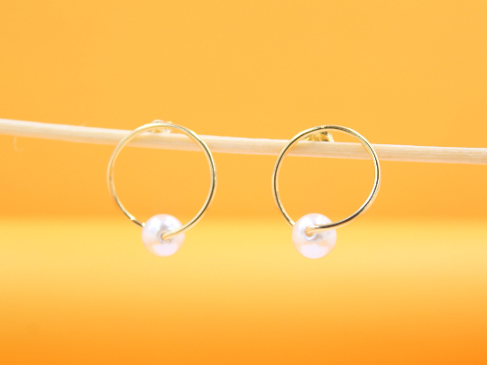 A pair of elegant gold hoop earrings featuring delicate pearl charms rests on a thin wooden stick against a warm orange background.