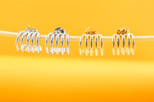Two pairs of multi-band hoop earrings in silver and gold rest on a thin wooden stick against a bright yellow background.