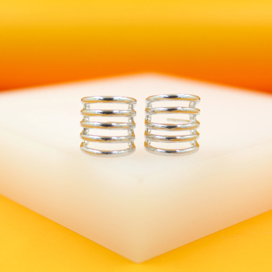 A pair of polished silver multi-band hoop earrings rests on a white display surface against a vibrant orange background.