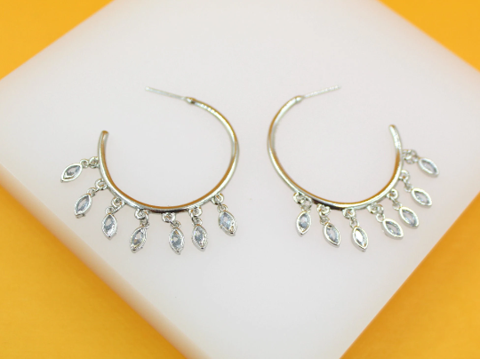 A pair of silver hoop earrings features dangling marquise-cut crystal charms against a white display surface.