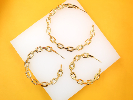 Three gold chain link hoop earrings of varying sizes lie arranged on a white display surface against a vibrant yellow background.