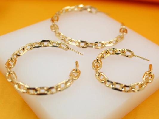 Three gold chain-link hoop earrings rest on a white display block against an orange background.