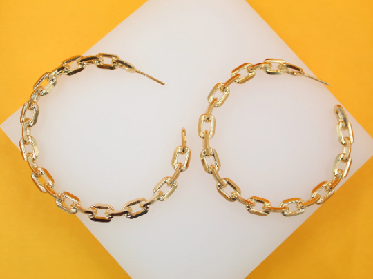 A pair of gold chain-link hoop earrings rests on a white display surface against a vibrant yellow background.