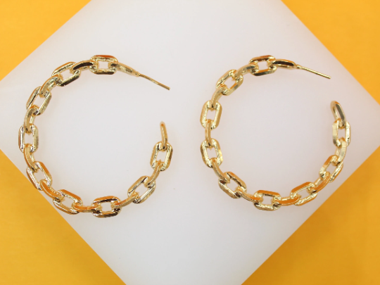 A pair of shiny gold chain link hoop earrings rest on a white display surface against a vibrant yellow background.