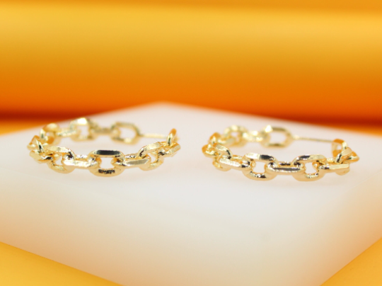 A pair of gold chain-link hoop earrings rests on a white display surface against a warm yellow background.