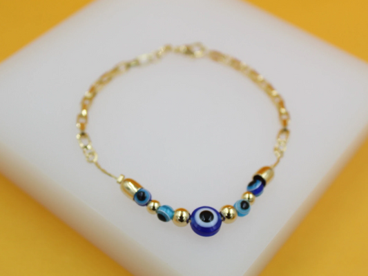 A delicate gold chain bracelet features a central blue evil eye charm flanked by smaller blue evil eye beads and gold spheres against a white surface.