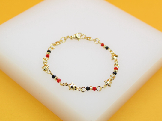 A delicate gold-plated children's bracelet features alternating red and black beads alongside tiny gold star charms, displayed on a white surface with a yellow background.