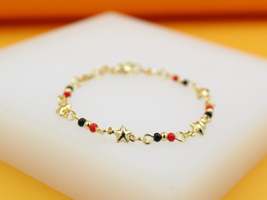 A delicate gold star bracelet rests on a white display surface, featuring alternating black and red beads along its chain.